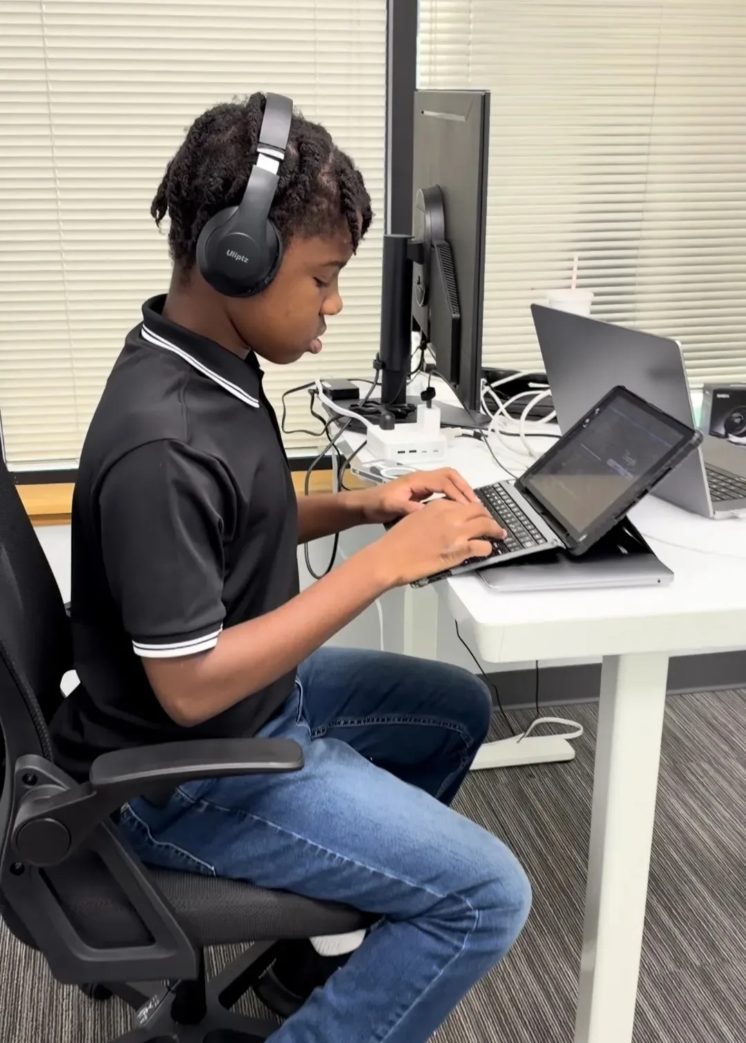 Titus, wearing headphones and a black polo, typing on a tablet with a keyboard at a white desk in an office with window blinds behind him.
